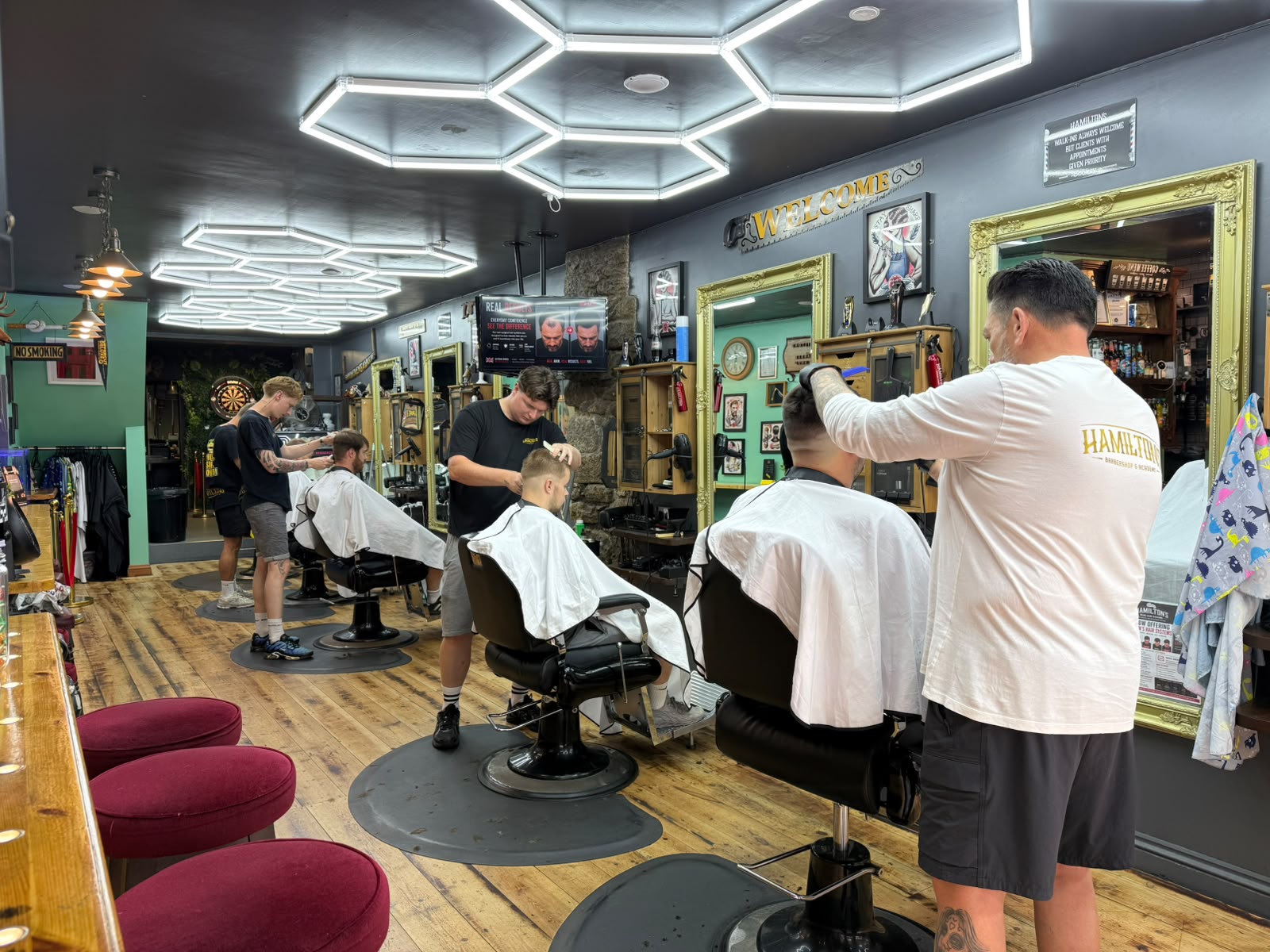 Inside Hamilton's Barbershop in Redruth — barbers cutting hair at three chairs beneath hexagon lighting, with the wooden coffee bar and gilt mirrors